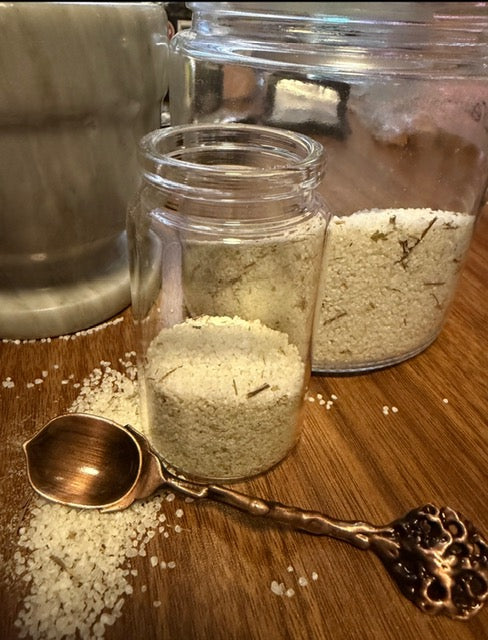 Green witch's salt blend in a clear glass jar with a wooden scoop and herbal pieces