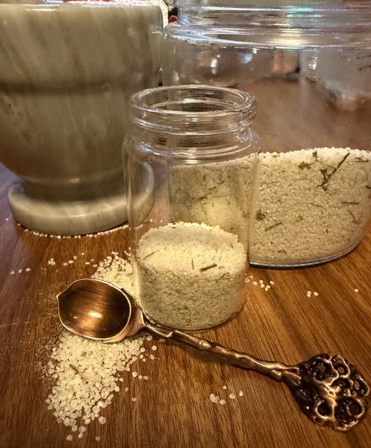 Green witch's salt blend in a clear glass jar with a wooden scoop and a larger jar behind it